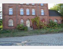 Backsteinbau mit drei Etagen und mehreren weißen Fenstern, dessen Fassade teilweise mit Efeu und rotem Weinlaub bewachsen ist. Vor dem Gebäude befinden sich eine kleine Treppe zum Eingang und ein unbefestigter Weg mit Kopfsteinpflaster.