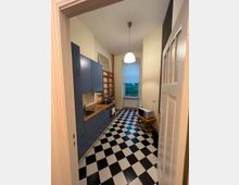 Kleine Küche mit blauen Schränken und einer Holzarbeitsplatte auf der linken Seite. Der Boden hat ein Schwarz-Weiß-Schachbrettmuster, und am Ende des Raumes befindet sich ein Fenster mit heruntergelassenem Rollo.