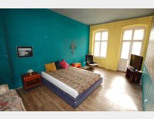 Schlafzimmer mit Holzboden, einer türkisfarbenen und einer gelben Wand. In der Mitte steht ein Doppelbett mit gemusterter Tagesdecke und bunten Kissen, flankiert von Nachttischen; ein Ledersessel, ein kleiner Fernseher auf einem Tisch und große Fenster sorgen für Licht.