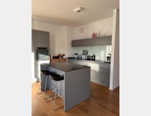 Moderne Küche mit grauen Einbauschränken und -schubladen, einer glänzenden Kücheninsel mit zwei schwarzen Barhockern und einer Rückwand aus Glas. Der Boden ist aus hellem Holz, die Wände sind weiß, und auf der Arbeitsfläche stehen Küchenutensilien und Geräte.