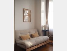 Bright living room corner with a beige tufted futon sofa on a wooden frame and two tan pillows, beneath a framed abstract print on a white wall. A tall window with sheer curtains and drapes lets in sunlight, with a small side table and pleated lamp beside the sofa on a wooden floor.