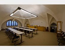 Ein Raum mit gotischen Gewölbedecken und großen, verzierten Fenstern. Der Raum ist mit Tischen, Stühlen und einem erhöhten Pult ausgestattet, vermutlich für Unterricht oder Versammlungen.