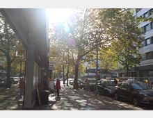 Straßenszene in einer Stadt mit Reihen von geparkten Autos, Fußgängern und mehreren Schildern, darunter BVG- und Lotto-Schilder. Im Hintergrund stehen moderne Gebäude, während Bäume mit herbstlichem Laub das Sonnenlicht filtern.