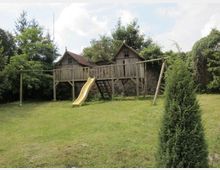Holz-Spielplatz mit zwei kleinen Häuschen auf Stelzen, einer gelben Rutsche und einer Leiter, umgeben von einer Rasenfläche und Bäumen im Hintergrund.