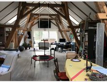 Großzügiger Dachraum mit sichtbaren Holzbalken und verschiedenen Wohnbereichen, darunter ein Esstisch mit gelber Tischdecke, eine Sitzecke, ein Trampolin und ein Arbeitsplatz mit Schreibtisch und Computer. Große Fenster lassen Tageslicht herein, und Regale mit Büchern sowie Dekorationen prägen den Raum.