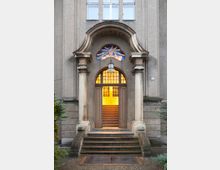 Eingang eines historischen Gebäudes mit zwei Säulen, einem gewölbten Türbogen und einem farbigen Bleiglasfenster mit religiösem Motiv. Die beleuchtete Tür führt zu einer Treppe, die ins Innere des Gebäudes führt, umgeben von einem gepflasterten Außenbereich mit Grünpflanzen.