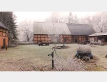 Ein verschneiter Bauernhof mit Fachwerkgebäuden, einem zentralen Scheunengebäude mit Wellblechdach, einem Handwasserpumpe in der Mitte und einem kleinen, mit Pflanzen gefüllten Karren im Vordergrund. Im Hintergrund sind blattlose Bäume und vereiste Äste zu sehen.