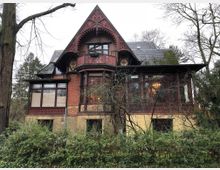 Historische Villa im Stil des Späthistorismus mit rotem Ziegelmauerwerk, detaillierten Holzschnitzereien am Giebel und einem überdachten Balkon. Umgeben von kahlen Bäumen und einem begrünten Vorgarten.