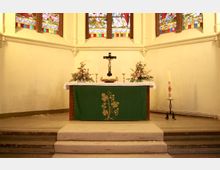 Altarraum einer Kirche mit einem Holzkreuz auf einem Tisch, der mit einem weißen Tuch und einem grünen Antependium mit Weinrebenmotiv bedeckt ist. Im Hintergrund sind bunte Glasfenster mit religiösen Darstellungen und seitlich Blumenarrangements sowie eine Osterkerze zu sehen.