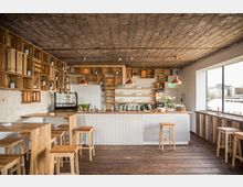 Gemütliches Café-Interieur mit Holzdekor, einer Theke aus weißgefrästem Holz, Holzregalen mit Dekorationen wie Pflanzen und Gläsern, sowie Hockern und Tischen aus hellem Holz. Große Fenster bieten Blick auf eine Uferlandschaft draußen.