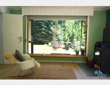 Wohnraum mit gemütlicher Einrichtung, bestehend aus einem Schaukelstuhl mit Fellüberwurf, einem Holzofen und einer großen Fensterfront mit Blick auf einen Garten mit Bäumen, einer Hängeschaukel und einer Picknickbank. Helle Holzböden und ein grünes Wanddesign dominieren den Raum.