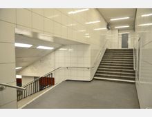 Treppenaufgang in einer U-Bahn-Station mit weißen gefliesten Wänden, Metallgeländern und Graffiti an den Wänden. Der Bereich ist hell beleuchtet und führt zu einer Tür im oberen Bereich.