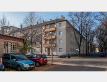 Wohngebäude aus der Nachkriegszeit mit hellbeiger Fassade und Balkonen an einer Straßenecke. Im Vordergrund sind parkende Autos, eine kleine Backsteinmauer mit einem Fenster sowie kahle Bäume an der Straße sichtbar.