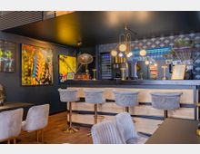 Elegante Bar mit moderner Einrichtung, einschließlich einer Theke mit hellen Holzvertäfelungen und grauen, gepolsterten Barhockern. An der dunkel gestrichenen Wand hängen bunte Kunstwerke, während goldene Lampen und Dekorationen wie Pfauenfedern und ein Grammophon den Raum schmücken.