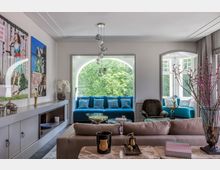 Wohnzimmer mit modernen Möbeln und großen Bögenfenstern, durch die man auf grüne Bäume blickt. Auffällige blaue Sitzbänke mit Kissen, ein Sofa im Vordergrund und ein Marmortisch mit Dekoobjekten bestimmen die Einrichtung; an der linken Wand sind Kunstwerke angebracht.