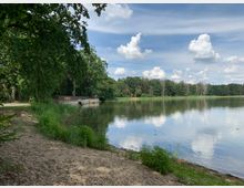 Naturlandschaft mit einem See, umgeben von dichtem Grün und einem Sandufer im Vordergrund. Am linken Ufer steht eine kleine Aussichtsplattform aus Holz, während der Himmel teils bewölkt ist und sich im Wasser spiegelt.