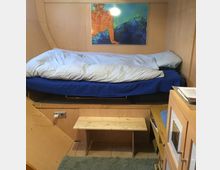 Holzvertäfelter Raum mit einer erhöhten Schlafnische, ausgestattet mit einer Matratze, blauer Bettwäsche und weißen Kissen. Unter der Schlafnische befinden sich ein kleines Holzregal und eine Steckdose, daneben steht ein Holzschemel; an der Wand hängt ein farbiges Kunstbild.