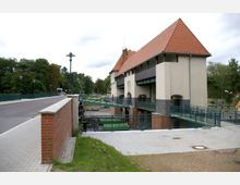 Brückenhaus einer historischen Schleusenanlage mit rotgedecktem Satteldach, hellem Putz und grünen Metallgeländern. Im Vordergrund sind ein Gehweg, eine Straßenbrücke und umgebende Grünflächen zu sehen, im Hintergrund befindet sich eine bewaldete Umgebung.