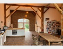 Großzügiger Wohnraum in einem renovierten Gebäude mit Holzbalkenkonstruktion, einer Küchenzeile links, einem langen Holztisch mit Stühlen rechts und einer großen Fensterfront mit Blick ins Freie. Im Hintergrund sind Bücherregale und Backsteinwände zu sehen.