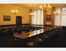 Ein Konferenzraum mit dunklen Holztischen und gepolsterten Stühlen, beleuchtet durch mehrere klassische Kronleuchter. Große Fenster mit halbtransparenten Vorhängen lassen Tageslicht in den Raum, während gerahmte Gemälde die Wände schmücken.