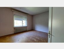 Leerer Raum mit hellen Wänden, einem großen Fenster mit Jalousien und einem Heizkörper darunter. Der Boden ist mit Fischgrätenparkett ausgelegt, und die Tür im Vordergrund ist leicht geöffnet.