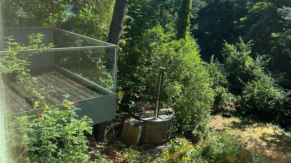 Terrasse aus Holz mit Glasgeländer an einem bewaldeten Hang, umgeben von dichtem Grün und hohen Bäumen. Unten im Gartenbereich sind ein kleiner gepflasterter Platz sowie eine runde Holzwanne mit aufrechtem Metallrohr zu sehen.