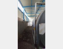 Innenansicht einer Lagerhalle mit Wellblechdach und blauen Stahlträgern. Rechts im Bild befindet sich ein großer, rostiger Metallbehälter, links eine Betonwand und ein Boden mit Holzspänen oder Schmutz.