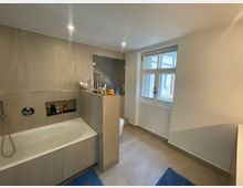 Modernes Badezimmer mit eingelassener Badewanne, grauen Wand- und Bodenfliesen sowie einer Nischenablage in der Wand. Ein großes Fenster sorgt für Tageslicht; daneben befinden sich eine halbhohe Trennwand, ein dunkler Duschbereich und ein WC.