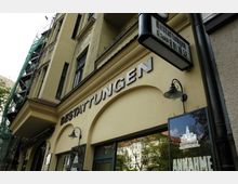 Fassade eines gelben Gebäudes mit einem Geschäft für Bestattungen, erkennbar durch den Schriftzug „BESTATTUNGEN“ und ein beleuchtetes Firmenschild. Im oberen Bereich sind mehrere Fenster und ein Teil eines eingerüsteten Fassadenteils sichtbar.