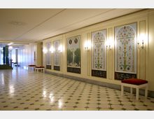 Heller, eleganter Innenraum mit dekorativen Wandmalereien, die florale und figürliche Muster zeigen, beleuchtet durch Wandlampen. Der Boden besteht aus sechseckigen Fliesen, und entlang der Wände stehen gepolsterte Bänke mit roten Sitzflächen.