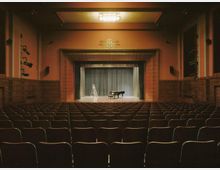 Innenraum eines Theatersaals mit leeren Sitzreihen, Blick auf eine Bühne mit einem geschlossenen Vorhang. Auf der Bühne stehen eine Leiter und ein schwarzer Flügel, umgeben von Holzvertäfelungen und seitlicher Beleuchtung.