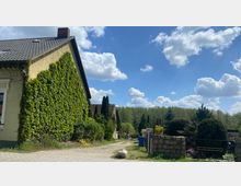 Ein Dorfweg mit einem gelben Haus, dessen Wand mit Efeu bewachsen ist. Im Hintergrund befinden sich Bäume, Hecken und weitere Gebäude, während der Himmel mit vereinzelten Wolken bedeckt ist.