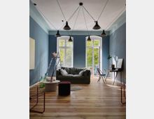 Wohnzimmer mit hellblauen Wänden und einem Holzboden. Zentral steht ein graues Sofa vor großen Doppelfenstern mit Blick ins Grüne, dazu ein kleiner Couchtisch, eine Stehleiter mit Kissen, ein Schreibtisch und mehrere Hängelampen von der Decke.