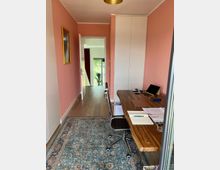 Ein kleiner Arbeitsbereich mit rosa gestrichenen Wänden, einem hölzernen Schreibtisch, einem schwarzen Bürostuhl und einem blauen Teppich mit orientalischem Muster. Im Hintergrund führt ein Flur mit Holzboden zu einem Raum mit großen Fenstern und Blick ins Grüne.