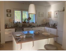 Eine moderne Wohnküche mit weißer Einrichtung und Holzarbeitsflächen. Im Mittelpunkt steht eine Kücheninsel mit Geschirr und Essensresten, im Hintergrund ein Fenster mit Pflanzen auf der Fensterbank sowie Küchenutensilien und Wanddekorationen.