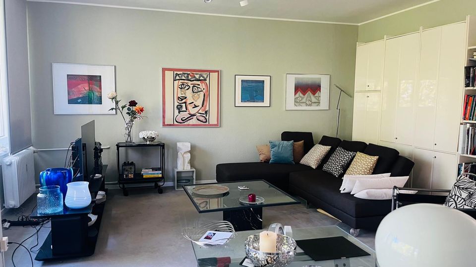 Modernes Wohnzimmer mit hellgrauen Wänden, großem schwarzen Ecksofa mit vielen Kissen und Glas-Couchtisch auf grauem Teppich. An der Wand hängen mehrere gerahmte Kunstbilder; links steht ein TV-Möbel vor einem Fenster, rechts sind hohe weiße Einbauschränke und ein Regal zu sehen.