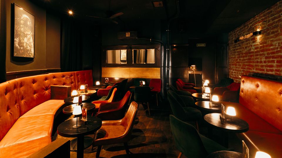 Dunkler Bar- oder Lounge-Innenraum mit orangefarbenen, gepolsterten Sitzbänken, runden schwarzen Tischen und kleinen Tischlampen. An der Seite ist eine freiliegende Backsteinwand, im Hintergrund eine Theke mit Spiegeln und gedämpfter Deckenbeleuchtung.