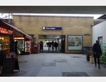 Eingang zur S-Bahn-Station "Rathaus Steglitz" mit gelben Backsteinwänden, einem blauen Stationsschild und einem überdachten Durchgang. Links ist ein kleiner Laden mit rotem Schild und Auslagen zu sehen, rechts eine Wand mit Plakaten und Personen im Vorbeigehen.
