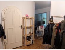 Ein kleiner Eingangsbereich mit hellem Holzboden, einer weißen, dekorativ verzierten Tür, einem Holzregal mit Ablagen und einer Kleiderstange mit Jacken. Im Hintergrund ist ein Raum mit einer Wand aus strukturiertem Glas erkennbar.