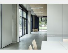 Moderner Innenraum eines Gebäudes mit großen Fensterfronten, die einen Blick auf eine Grünfläche ermöglichen. Der Raum hat eine minimalistische Einrichtung mit Holzstühlen, einem hellen Tisch und Betonwänden, sowie abgehängte Decken mit eingebauten Leuchten.