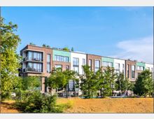 Modernes Wohngebäude mit einer Fassade aus Backstein und hellen Elementen, großen Fensterflächen und Balkonen, umgeben von Bäumen und Grünflächen, unter einem klaren blauen Himmel.