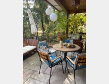 Eine überdachte Terrasse mit Holzmöbeln, darunter ein runder Tisch und Stühle mit Kissen, umgeben von üppiger Vegetation. Im Hintergrund sind Laternen und ein Sonnenschirm sichtbar, während Pflanzen und Dekorationen eine gemütliche Atmosphäre schaffen.