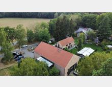 Luftaufnahme eines kleinen Landguts mit mehreren Backsteingebäuden mit roten Ziegeldächern, umgeben von Bäumen und einer Wiese. Im Vordergrund ein großes Hauptgebäude, dahinter weitere Gebäude, ein Parkplatz und Sitzbereiche im Freien.