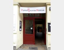 Eingangsbereich des Puppentheater-Museums mit einer roten Tür und einem weißen Banner darüber, auf dem der Schriftzug „Puppentheater-Museum“ steht. Der Eingang führt durch einen überdachten Durchgang mit Blick auf einen Innenhof.