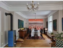 Esszimmer in einem klassischen Wohnraum mit Holzboden, einem Esstisch mit weißen Tischdecken und schwarzen Stühlen. Links ein blauer Kaminofen, rechts große Fenster mit viel Tageslicht; an der Stirnseite eine auffällige Wandmalerei mit roter Landschaft.