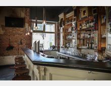 Gemütliche Bar mit Ziegelsteinwänden, einer langen Theke aus dunklem Holz und mehreren Barhockern. Im Hintergrund befinden sich beleuchtete Regale, die mit verschiedenen Flaschen und Gläsern bestückt sind, sowie ein großes Fenster, das Tageslicht hereinlässt.