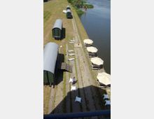 Mehrere grüne, historische Eisenbahnwaggons stehen auf einem begrünten Gelände mit gepflasterten Wegen. Daneben befindet sich ein Uferbereich mit Sonnenliegen und beigefarbenen Sonnenschirmen direkt am Wasser.