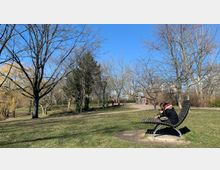 Ein Park mit kahlen Bäumen im Winter oder frühen Frühling, einigen Wiesenflächen und geschwungenen Spazierwegen. Auf der rechten Seite sitzt eine Person in einer warmen Jacke auf einer gebogenen Holzbank, im Hintergrund sind Spielgeräte und weitere Parkbesucher zu sehen.