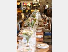 Langer, gedeckter Holztisch in einem rustikalen Restaurant mit unterschiedlichen Stühlen; dekoriert mit Blumenarrangements, Geschirr und Gläsern. Im Hintergrund eine Bar und unscharfe Details des gemütlichen Innenraums.