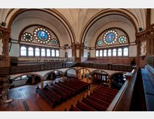 Innenraum einer Kirche mit roten Backsteinwänden, Rundbögen und Gewölbedecke. Die Ausstattung umfasst Holzgestühl, verzierte Emporen, bunte Glasfenster in Blumenmotiven sowie Statuen entlang der Wände.